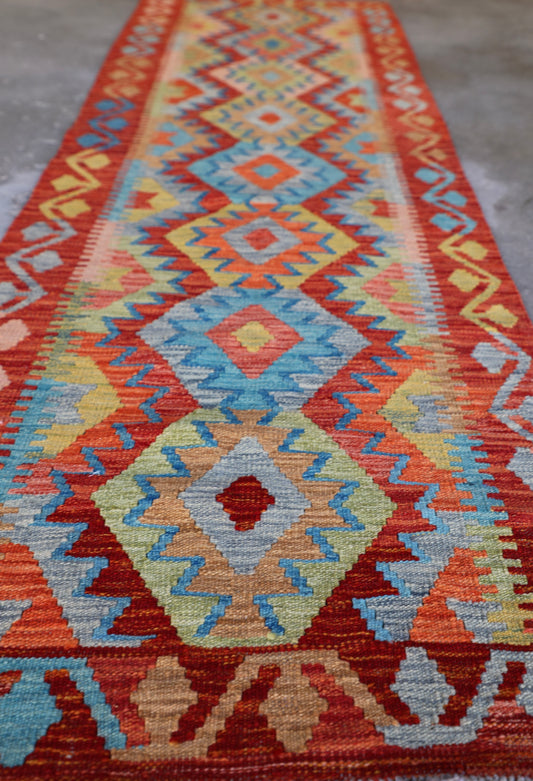 The Afghan Hand Woven Khyber Kilim Runner - 81 X 283. Crafted using traditional vegetable dyed wool, and featuring traditional geometric designs. Colours, reds, oranges, yellows and teal greens. Measures 81 wide X 283 long. Angle.