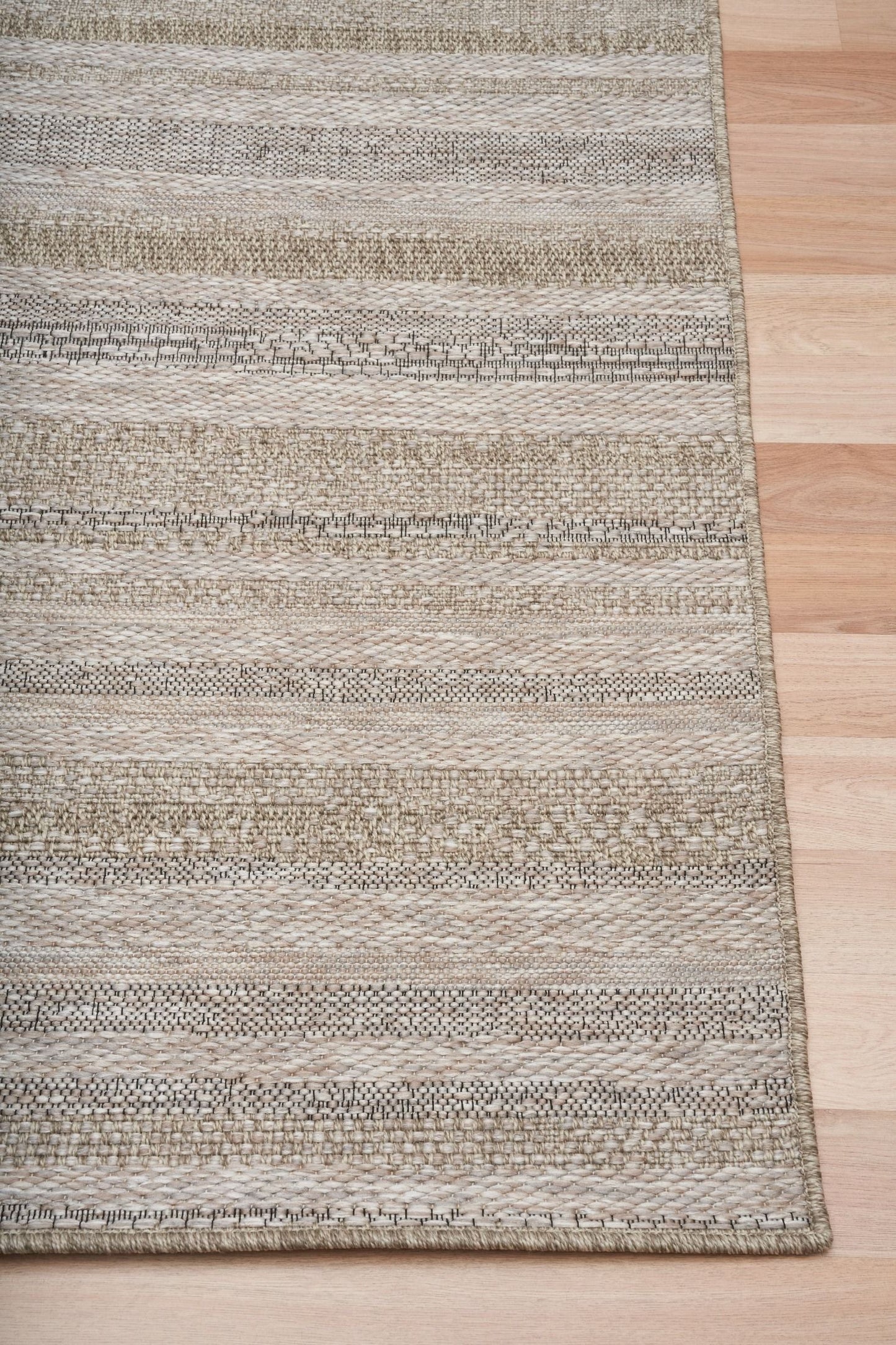 The Patio Linear Straw indoor/outdoor rug featuring neutral tones and a subtle textured pattern, power-loomed from UV-stabilised polypropylene for lasting durability. Colour Browns, Beiges and hints of black. 3 sizes. Corner.