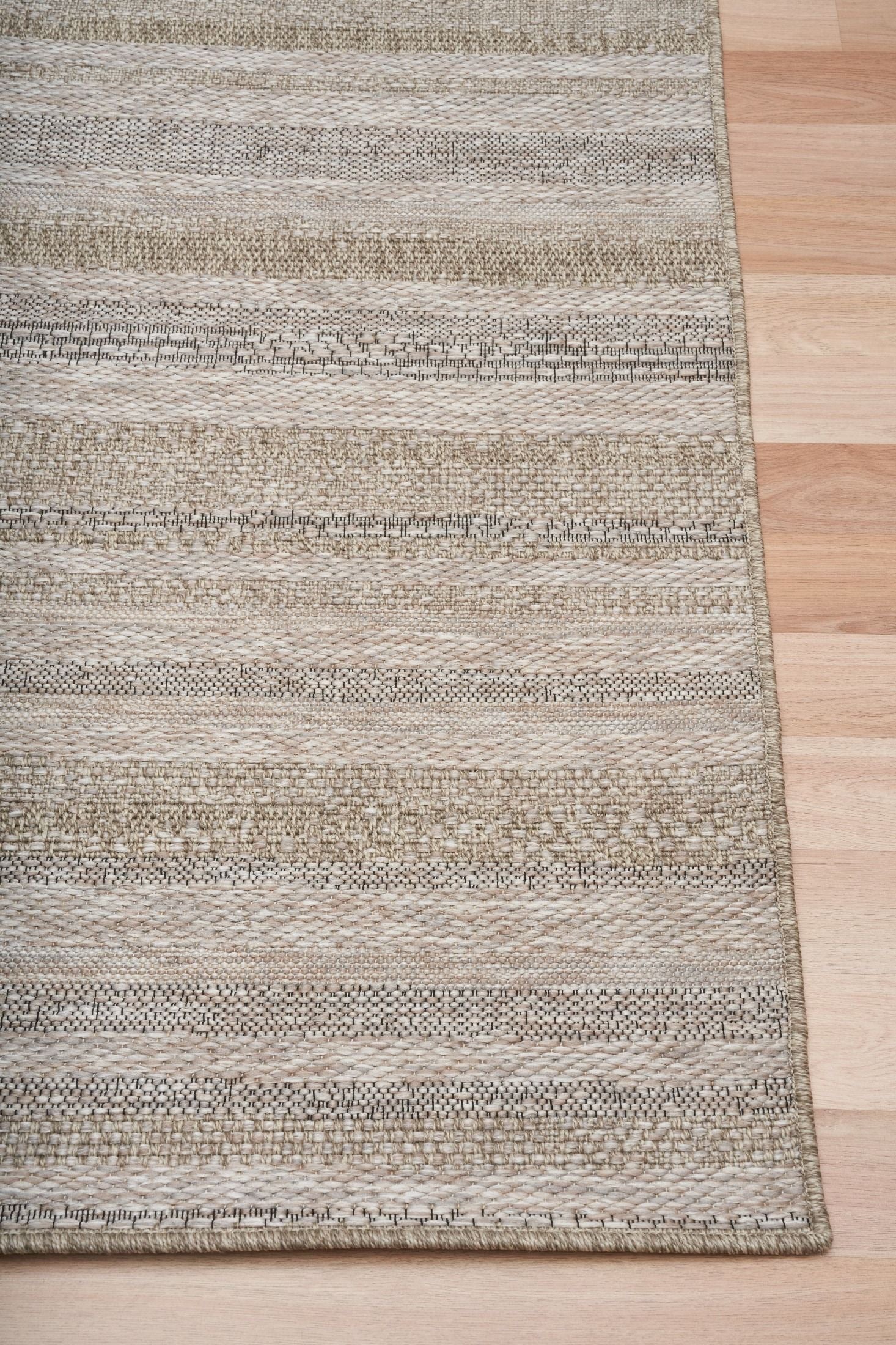 The Patio Linear Straw indoor/outdoor rug featuring neutral tones and a subtle textured pattern, power-loomed from UV-stabilised polypropylene for lasting durability. Colour Browns, Beiges and hints of black. 3 sizes. Corner.