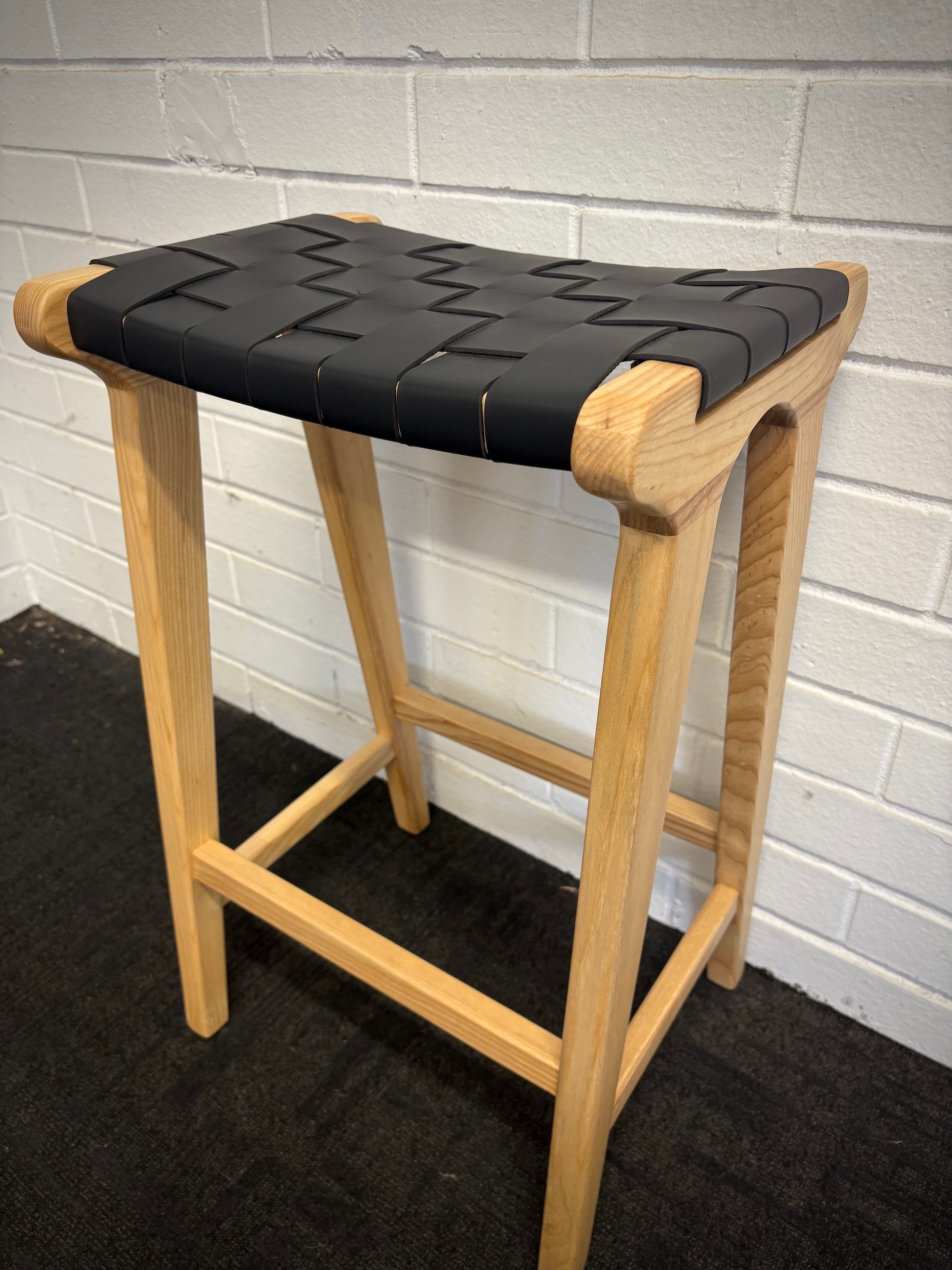The Thames Barstool, black leather, is designed with a contemporary style. Featuring a sustainable ash frame and a luxurious strapped saddle leather seat. Colour natural frame with black leather. Measures 410 wide X 320 depth X 640 height. Angle.